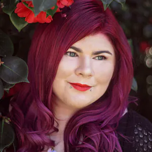 Zoe has dark red hair and is wearing red lipstick. She is standing in front of a bush with red flowers.
