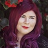 Zoe has dark red hair and is wearing red lipstick. She is standing in front of a bush with red flowers.