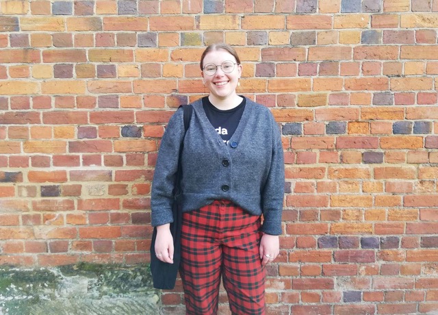 Amelia Saward wears a grey cardigan and red tartan pants and smiles while standing in front of a brick wall. 