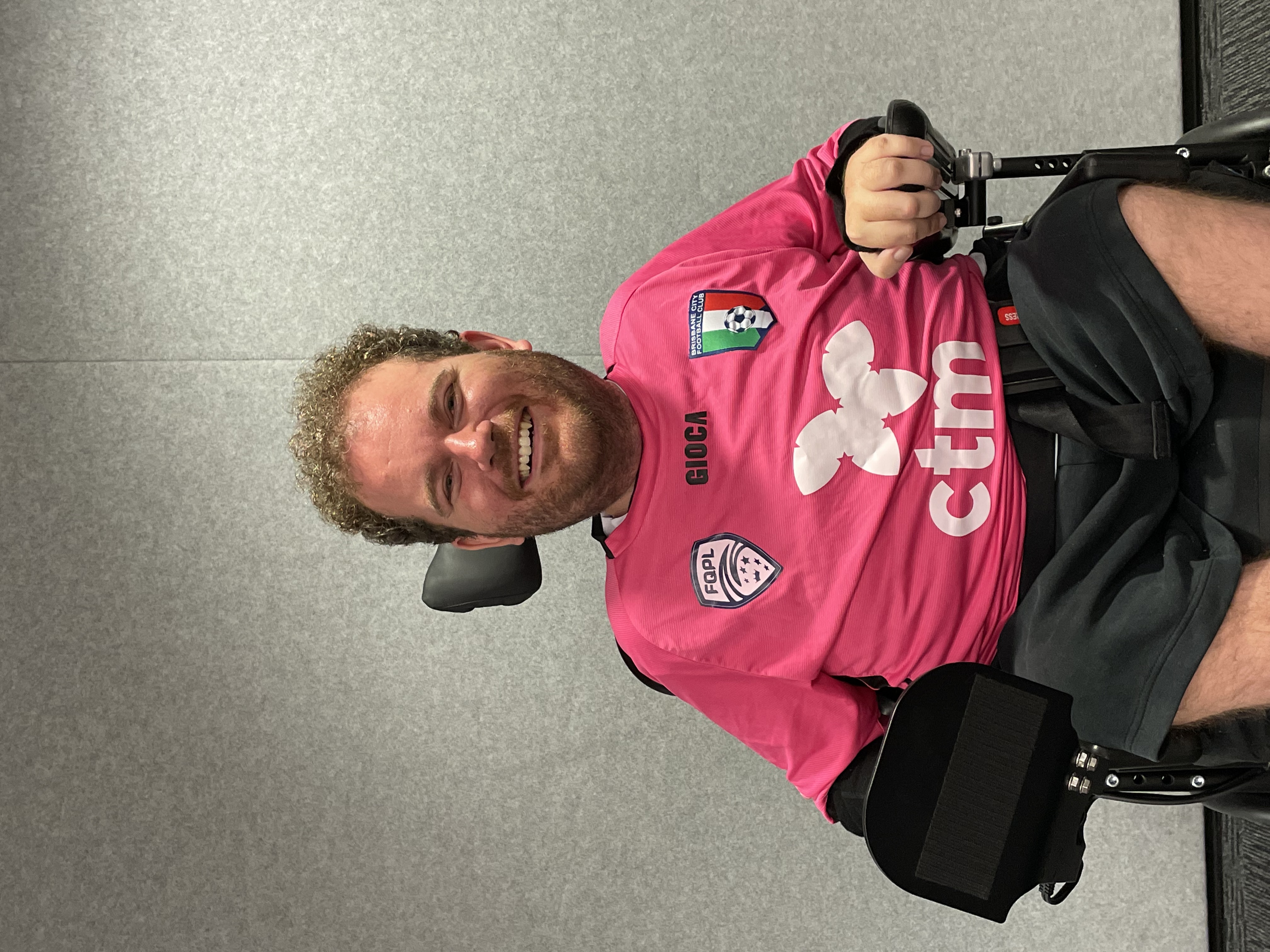 Tristram has curly brown hair, a beard and is smiling. He is wearing a pink football jersey and black shorts. Tristram uses a wheelchair. 