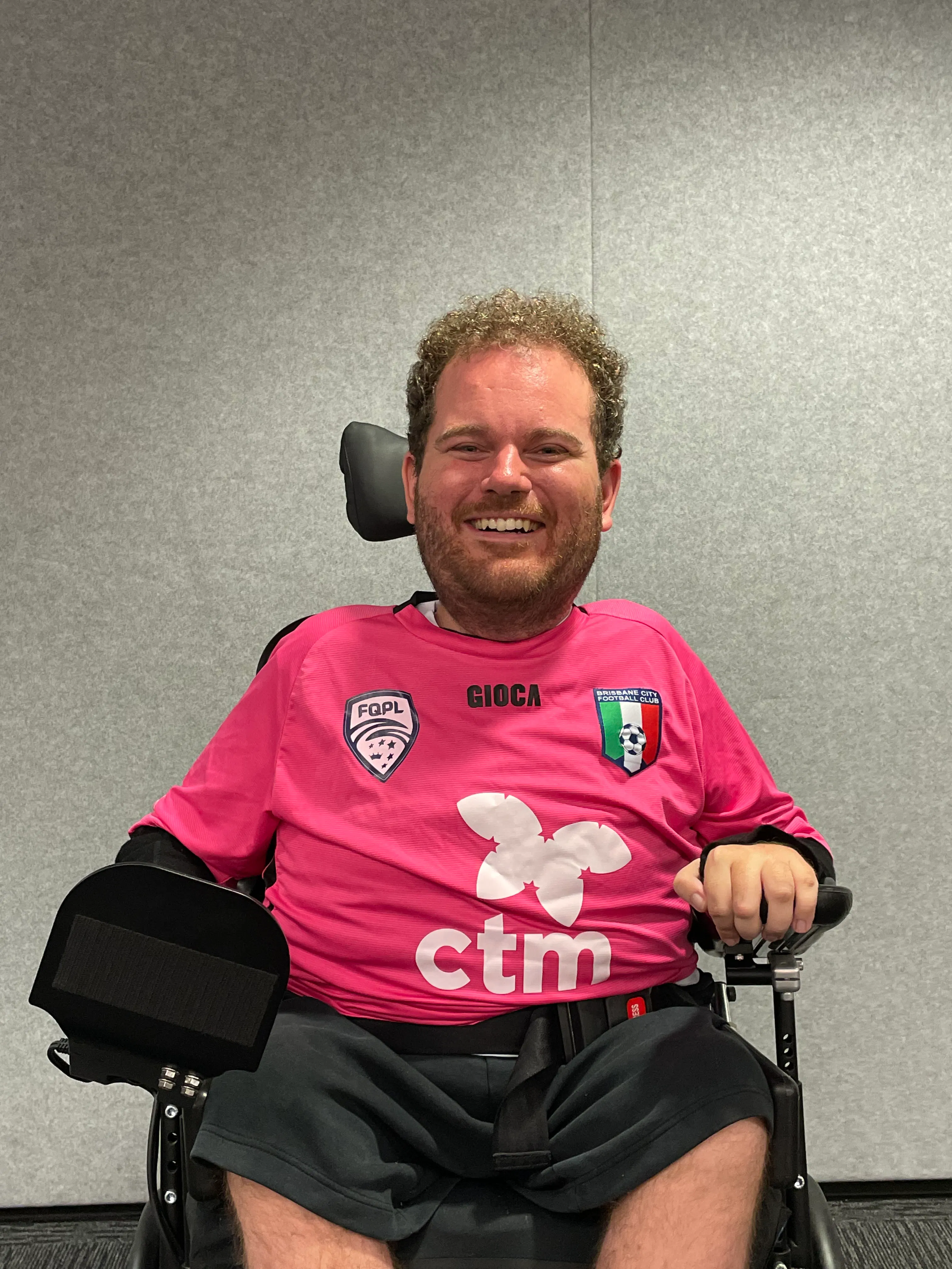 Tristram has curly brown hair, a beard and is smiling. He is wearing a pink football jersey and black shorts. Tristram uses a wheelchair. 