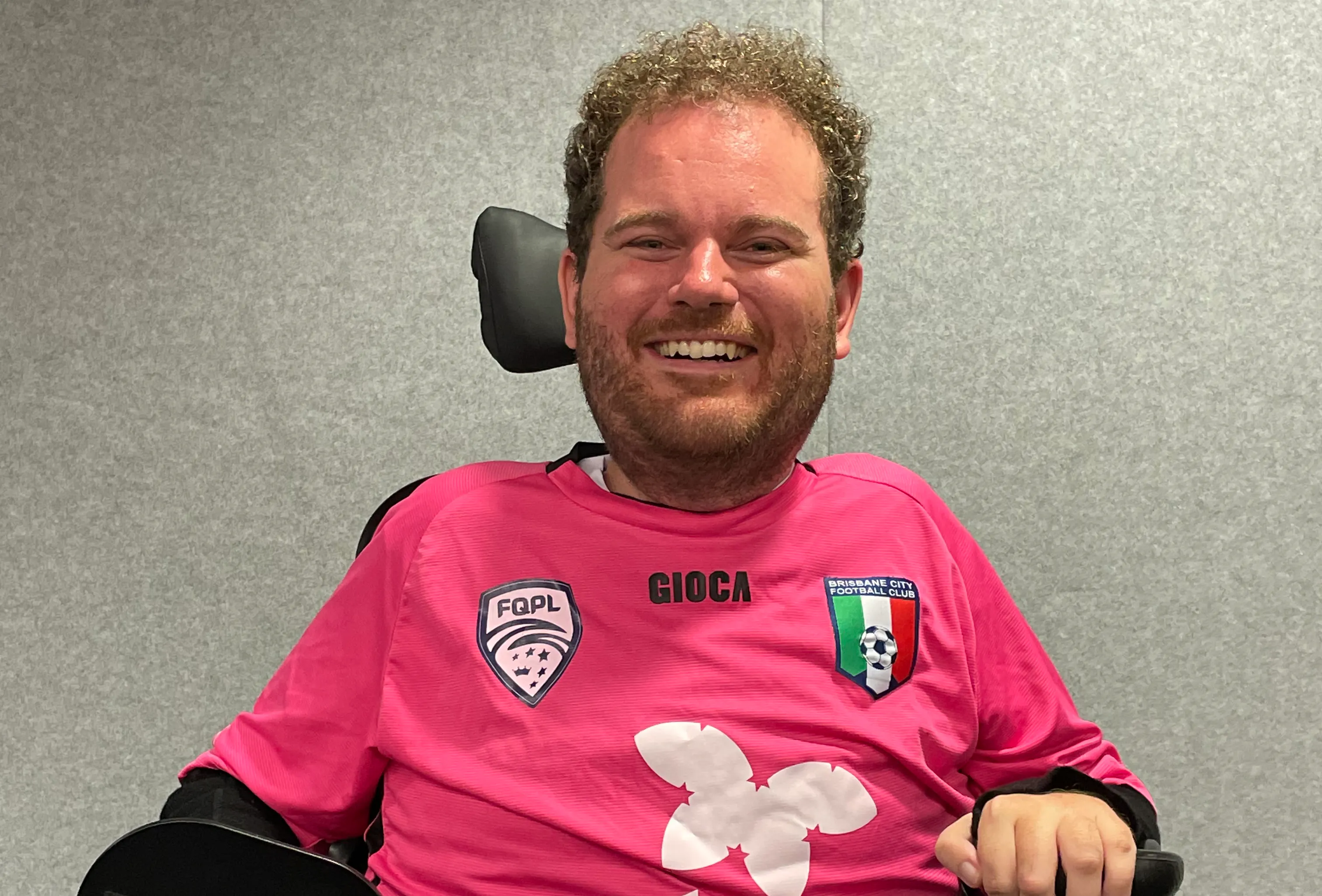 Tristram has curly brown hair, a beard and is smiling. He is wearing a pink football jersey and black shorts. Tristram uses a wheelchair.