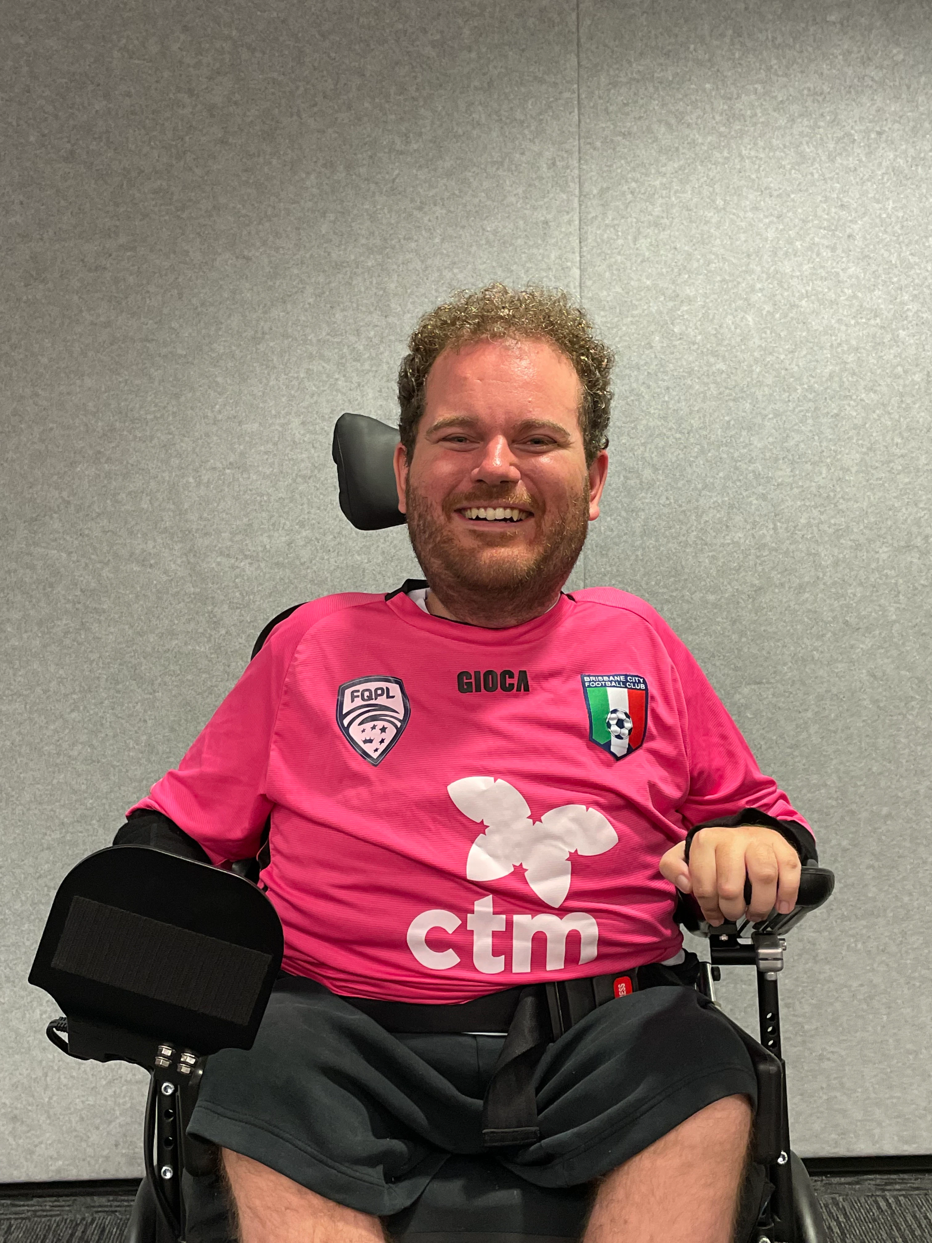 Tristram has curly brown hair, a beard and is smiling. He is wearing a pink football jersey and black shorts. Tristram uses a wheelchair.