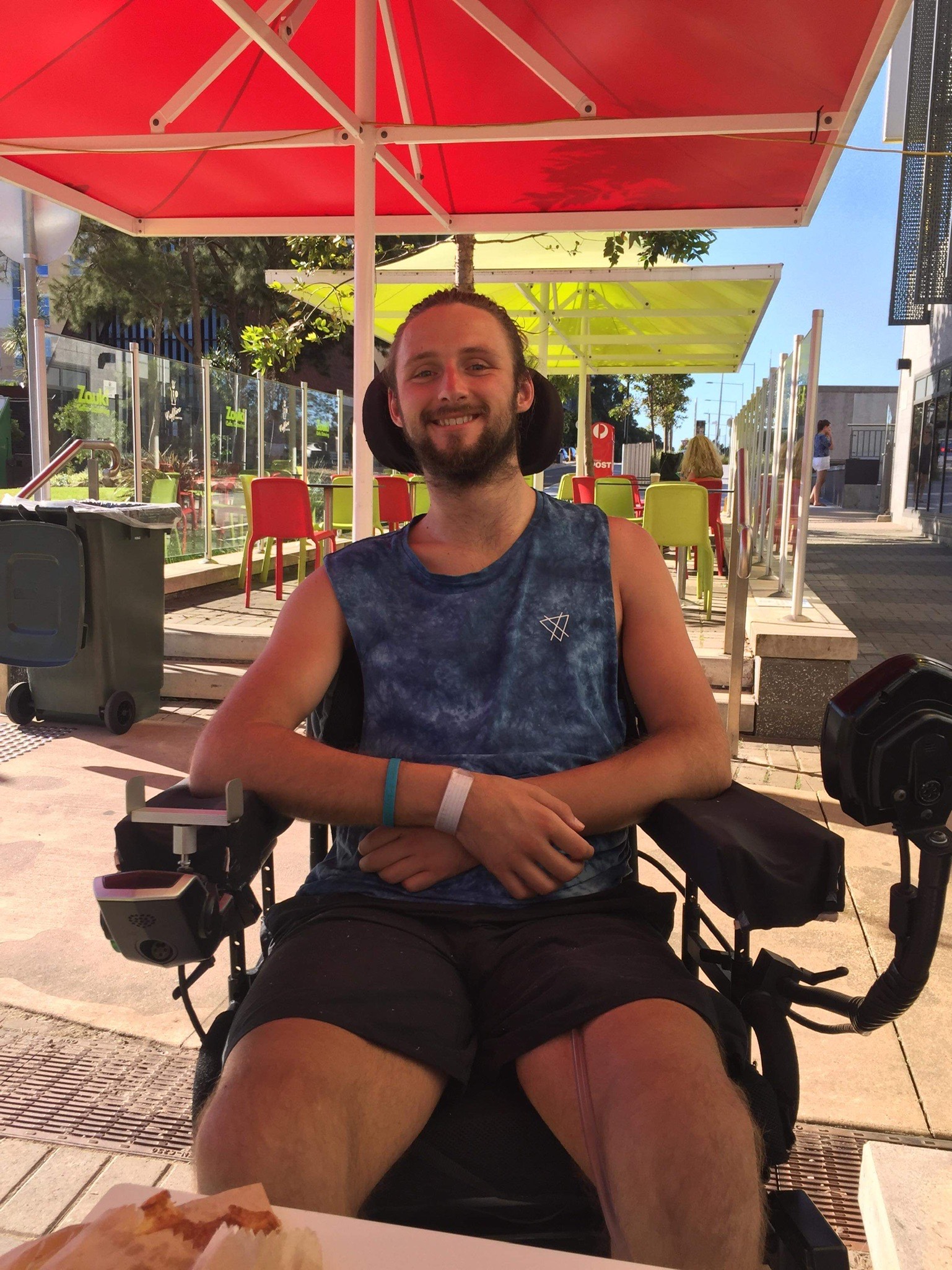 Steven is wearing a singlet shirt and shorts. He smiles widely with his arms folded in front of him. Red and green umbrellas are providing shade in the outdoor cafe. Steven in a wheelchair user. 