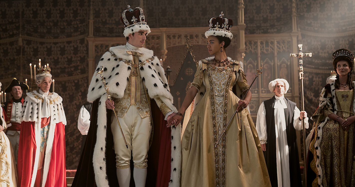 The characters of King George III and Queen Charlotte stand looking at one another in regal dress. 