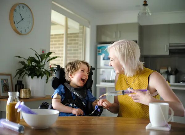 Arlo sits at the kitchen table with his support worker Maddie. They're smiling and laughing as he gets tube fed.
