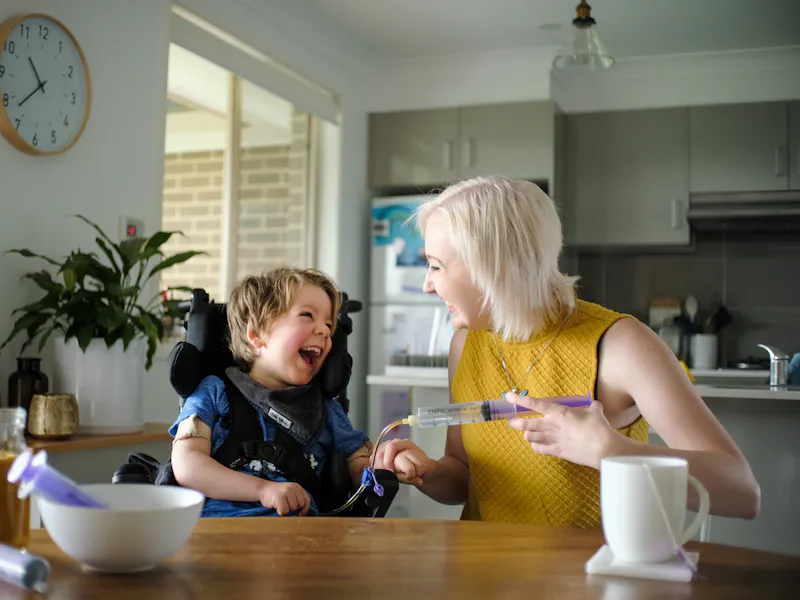 Arlo sits at the kitchen table with his support worker Maddie. They're smiling and laughing as he gets tube fed.
