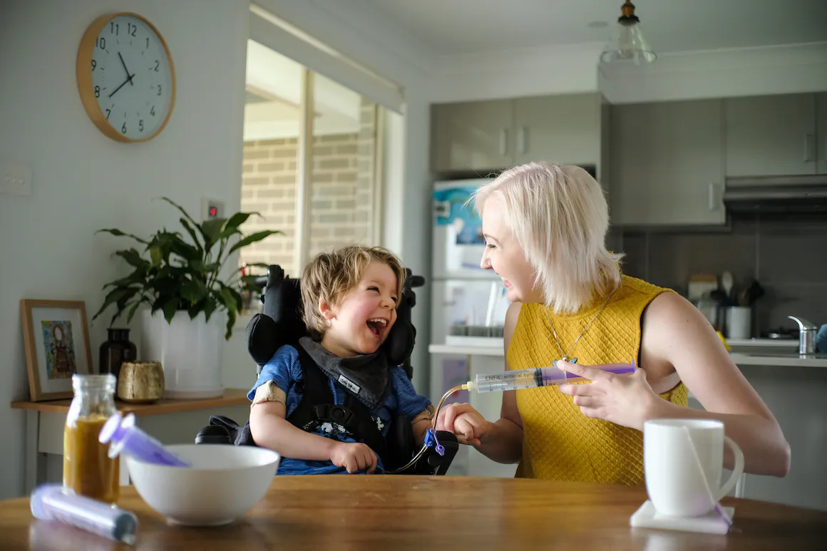 Arlo sits at the kitchen table with his support worker Maddie. They're smiling and laughing as he gets tube fed.