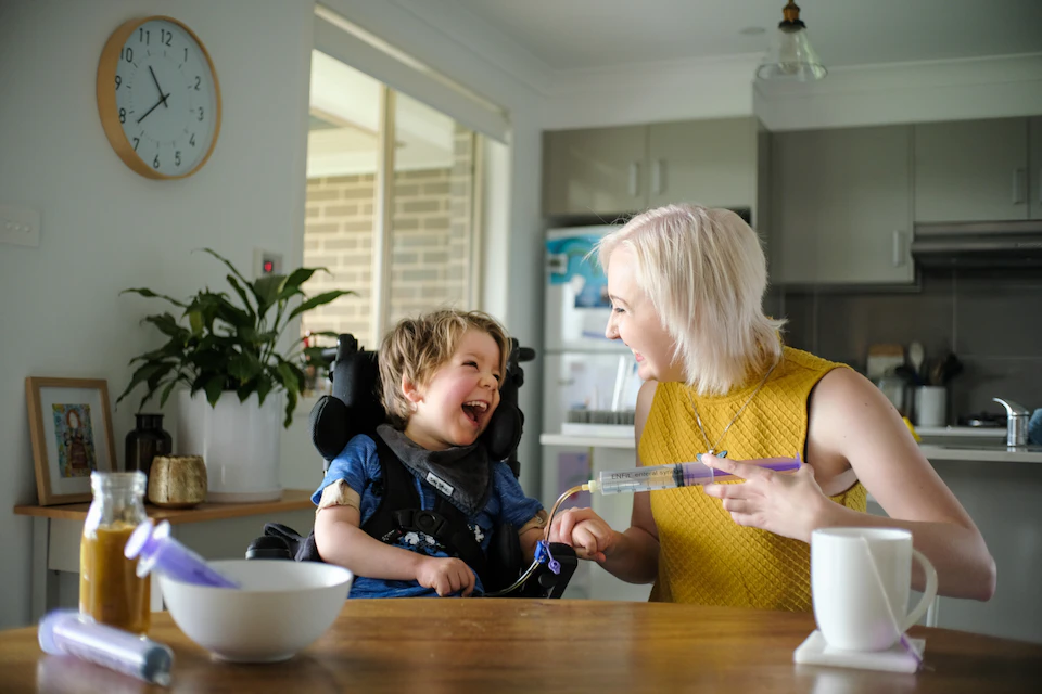 Arlo sits at the kitchen table with his support worker Maddie. They're smiling and laughing as he gets tube fed.