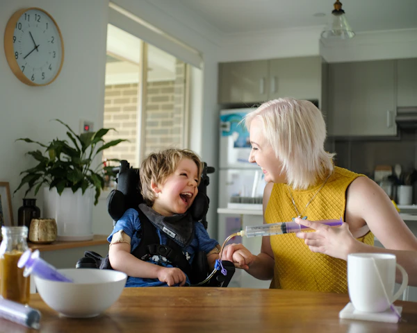Arlo sits at the kitchen table with his support worker Maddie. They're smiling and laughing as he gets tube fed.