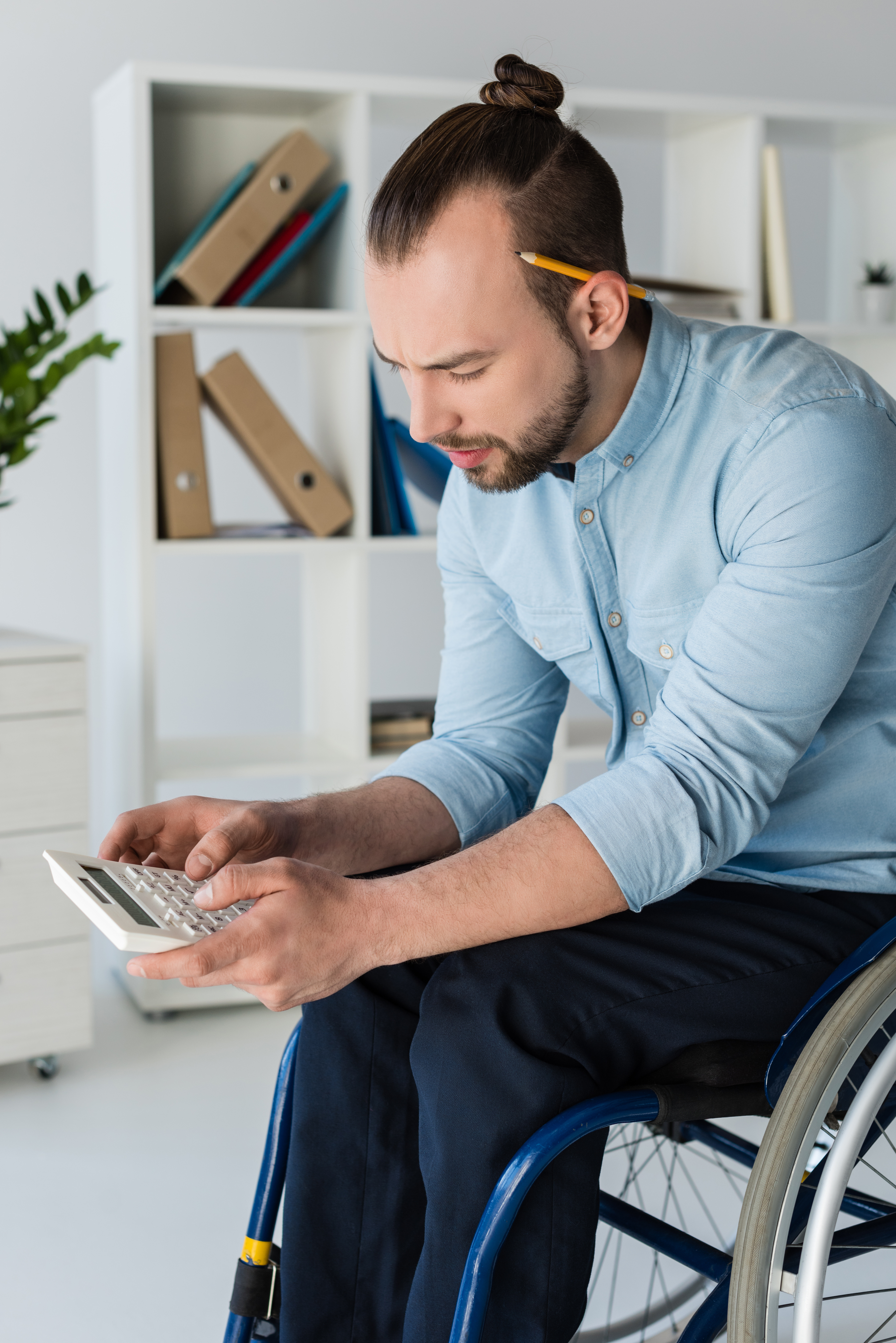 A man with dark hair tied in a small bun and a short beard is using a calculator. He is wearing a tight, light blue collared shirt and navy trousers. He has a yellow pencil behind his ear and is a wheelchair user. There are brown binders on white shelves in the background.