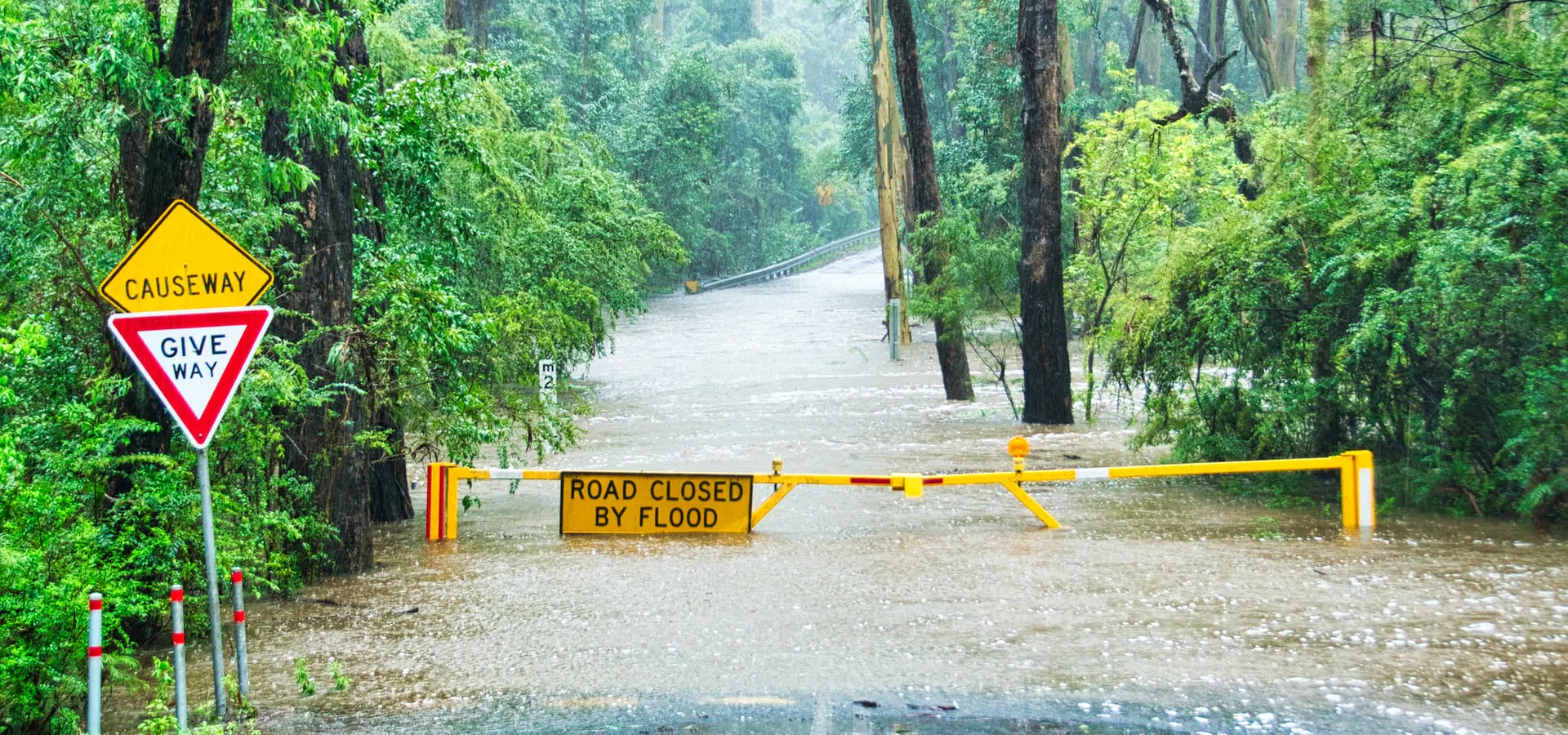Image shows: flooded road with "road closed by flood sign" across it