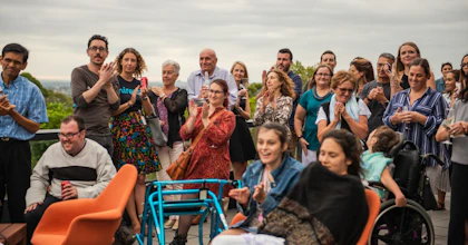 A group of the Hireup office support team stand and clap on a balcony outside. There are two people in wheelchairs in the foreground.