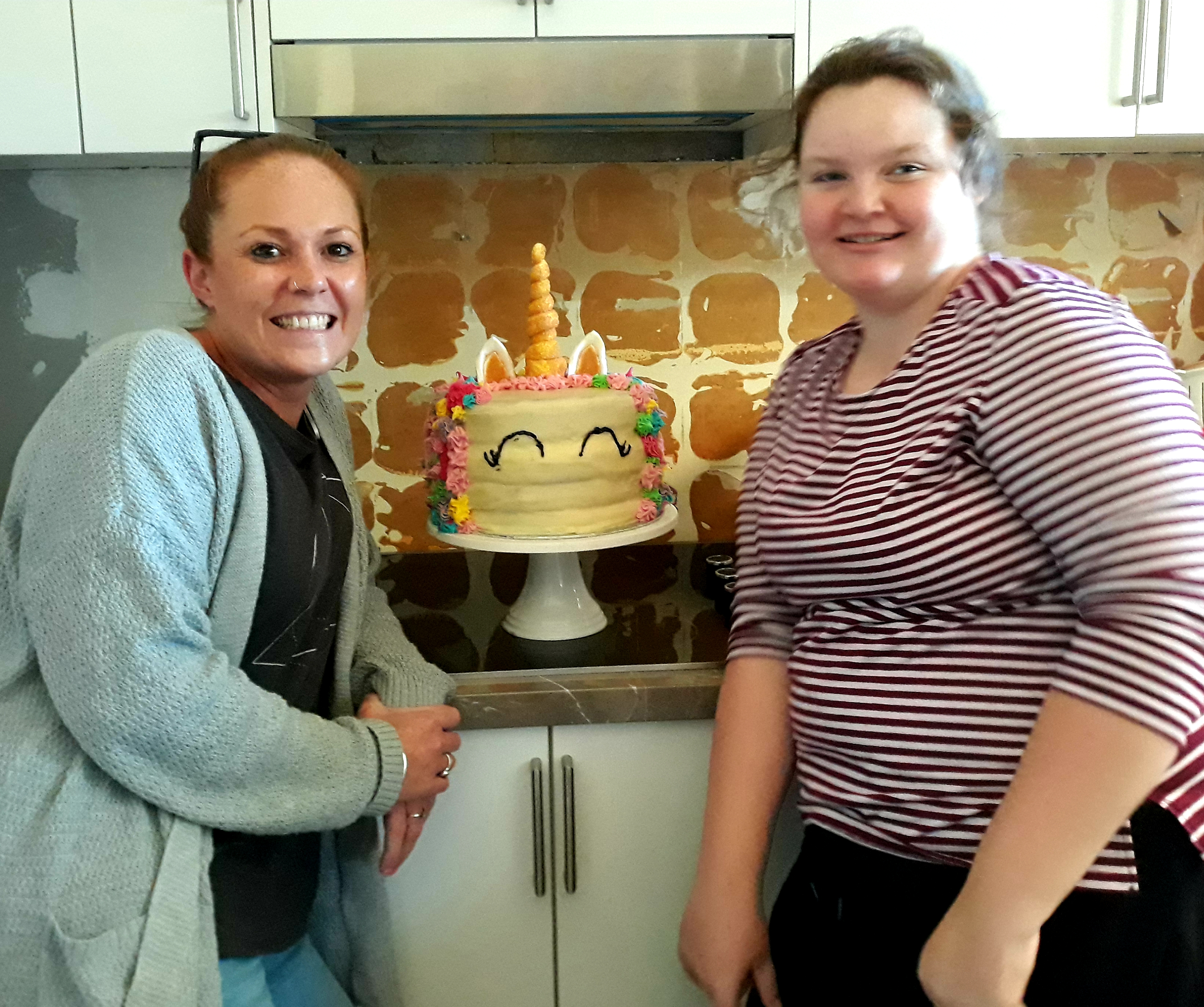 Claudia and Jasmin are standing in front of a birthday cake, smiling. Claudia has tied back red hair and is wearing a light blue cardigan over a grey shirt. Jasmin has brown hair and is wearing a red and white striped top.