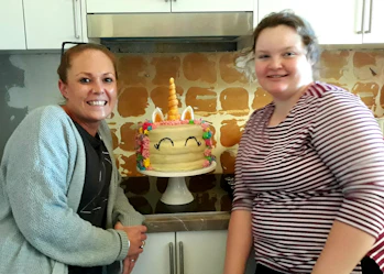 Claudia and Jasmin are standing in front of a birthday cake, smiling. Claudia has tied back red hair and is wearing a light blue cardigan over a grey shirt. Jasmin has brown hair and is wearing a red and white striped top.
