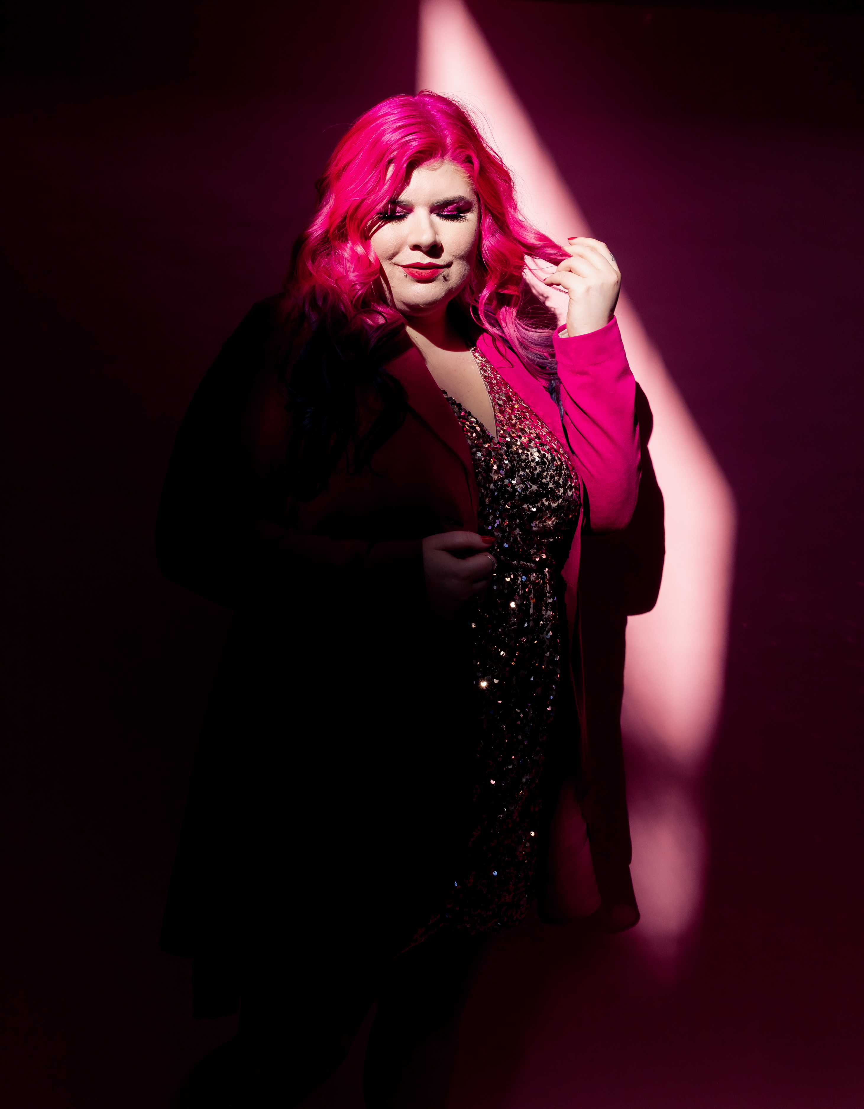 Disability advocate Zoe Simmons has long pink hair and wears a pink blazer over a glittering beaded dress. She stands against a pink backdrop in a beam of light. Photo by Emma Veness