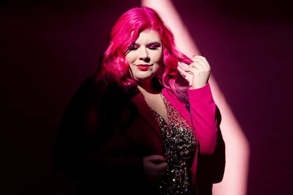 Disability advocate Zoe Simmons has long pink hair and wears a pink blazer over a glittering beaded dress. She stands against a pink backdrop in a beam of light. Photo by Emma Veness