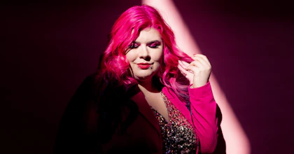 Disability advocate Zoe Simmons has long pink hair and wears a pink blazer over a glittering beaded dress. She stands against a pink backdrop in a beam of light. Photo by Emma Veness