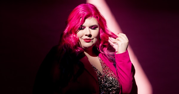 Disability advocate Zoe Simmons has long pink hair and wears a pink blazer over a glittering beaded dress. She stands against a pink backdrop in a beam of light. Photo by Emma Veness