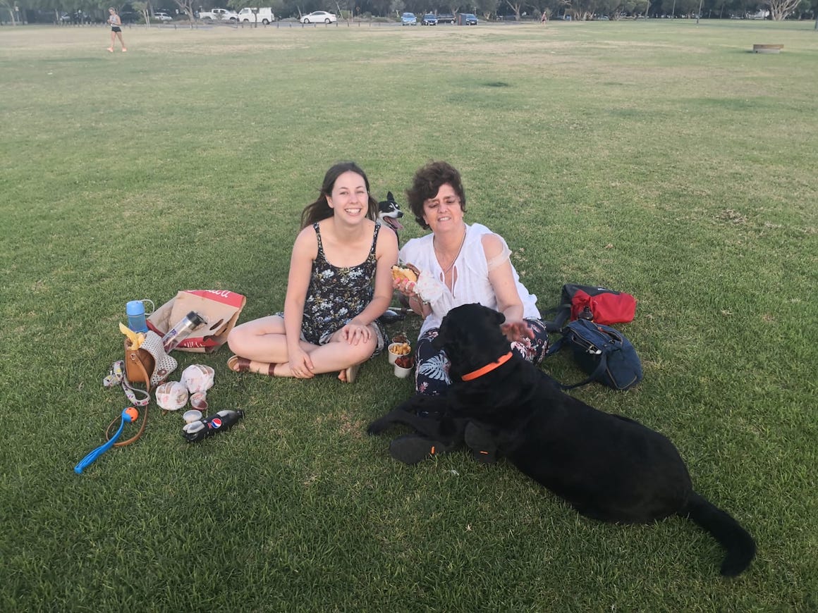 Tabitha and Gisele are sitting on the grass with her Guide Dog D'artagnan. They're enjoying a picnic by the Perth foreshore. 
