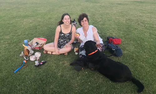 Tabitha and Gisele are sitting on the grass with her Guide Dog D'artagnan. They're enjoying a picnic by the Perth foreshore.