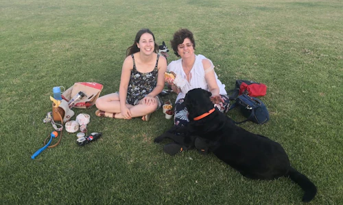 Tabitha and Gisele are sitting on the grass with her Guide Dog D'artagnan. They're enjoying a picnic by the Perth foreshore.