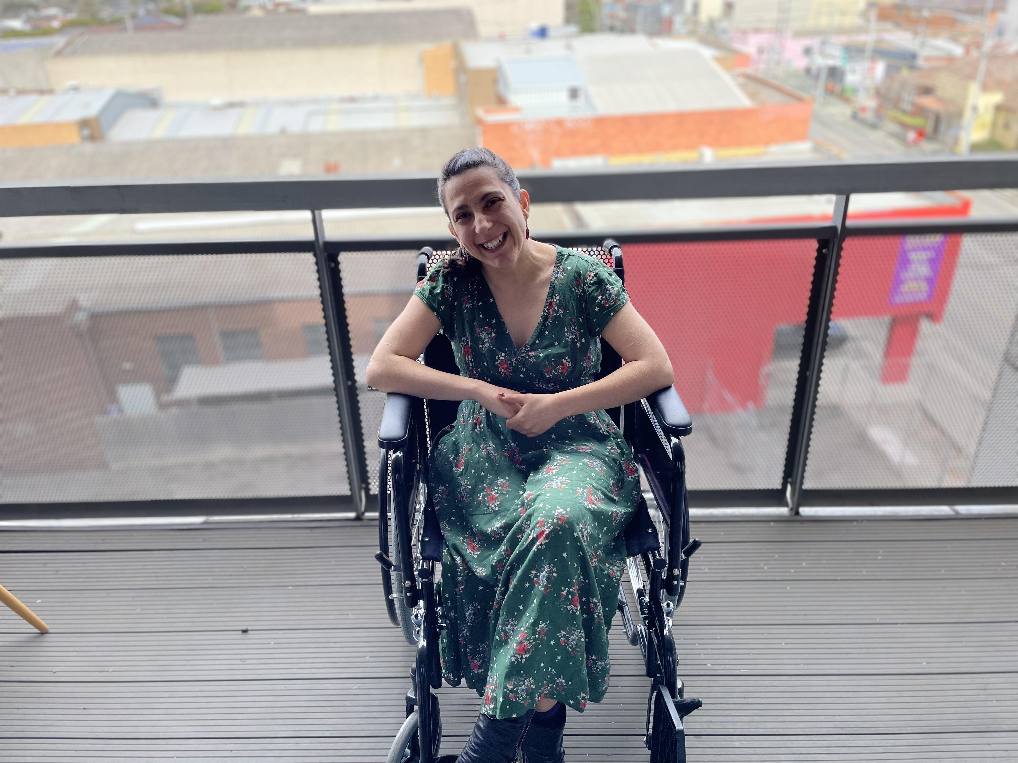 Writer Laura Pettenuzzo wears a green dress with small red and white flowers. Her hands are clasped and she's smiling. Laura is a wheelchair user. 