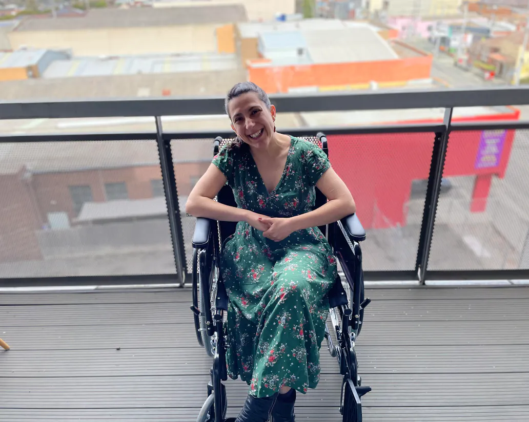 Writer Laura Pettenuzzo wears a green dress with small red and white flowers. Her hands are clasped and she's smiling. Laura is a wheelchair user.