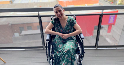Writer Laura Pettenuzzo wears a green dress with small red and white flowers. Her hands are clasped and she's smiling. Laura is a wheelchair user.