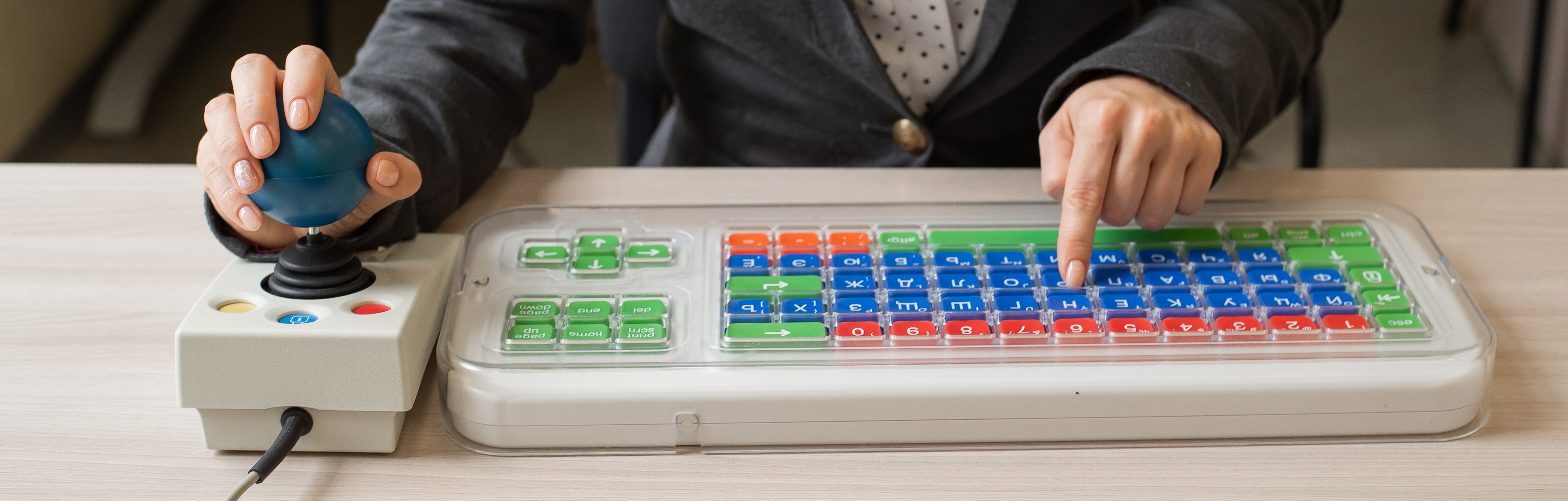 A person's hands are using a specialised computer mouse and keyboard. The mouse large circular blue joystick and the keyboard has bright green, blue and red keys with white symbols.
