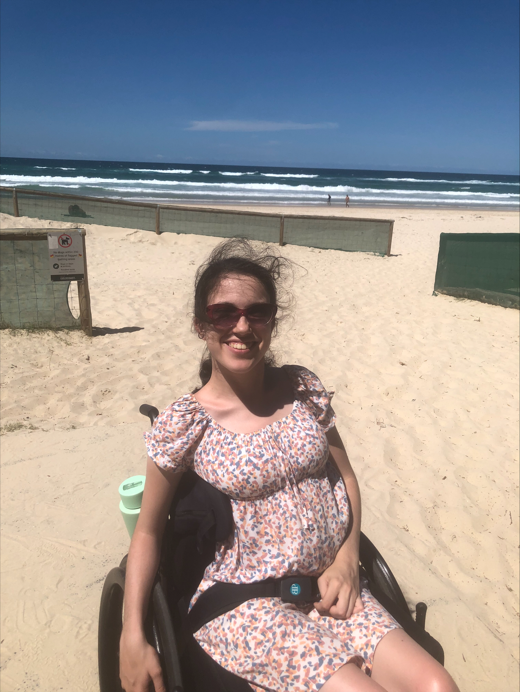 Disability advocate Hannah Diviney sits smiling at the beach in her wheelchair. She has her long dark hair tied back and wears sunglasses and a floral sundress. 