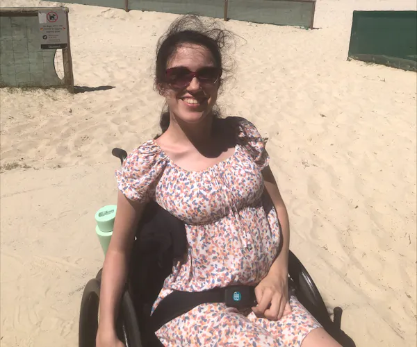 Disability advocate Hannah Diviney sits smiling at the beach in her wheelchair. She has her long dark hair tied back and wears sunglasses and a floral sundress.