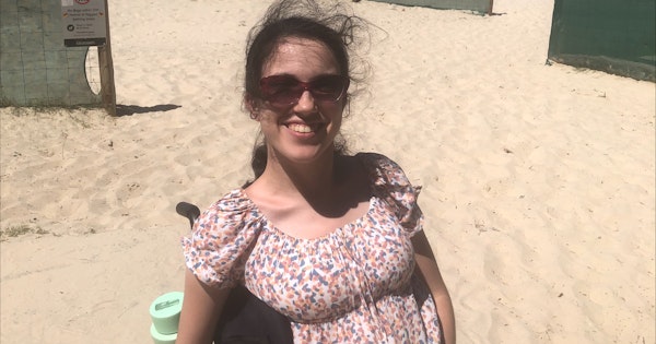 Disability advocate Hannah Diviney sits smiling at the beach in her wheelchair. She has her long dark hair tied back and wears sunglasses and a floral sundress.