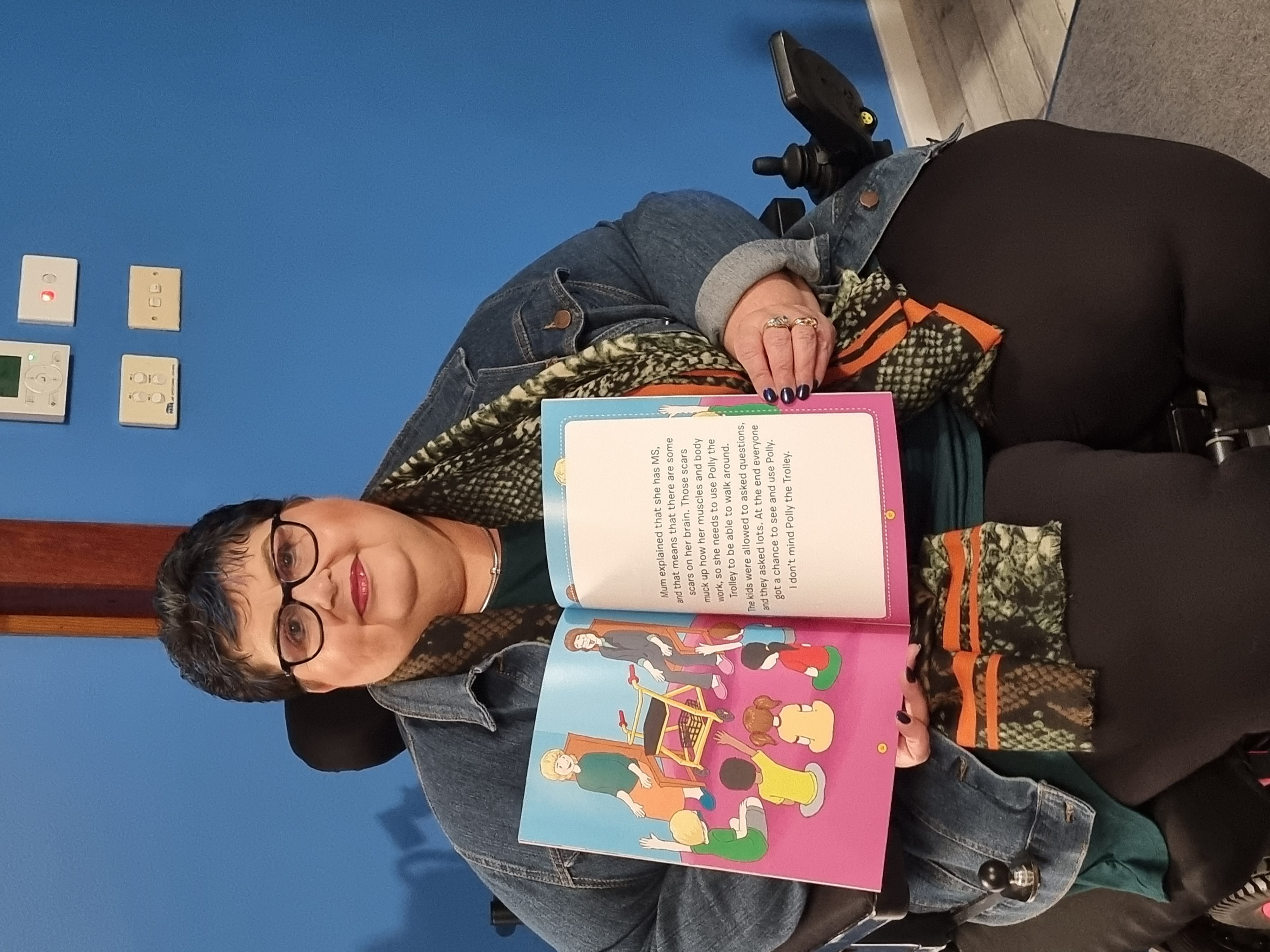 Leisa is sitting and holding a copy of her book, Polly the Trolley. She is wearing long black pants, a blue denim jacket and a scarf. Leisa has short dark hair and wears black rimmed glasses.