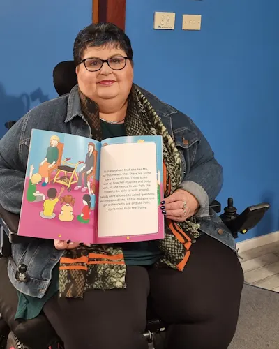 Leisa is sitting and holding a copy of her book, Polly the Trolley. She is wearing long black pants, a blue denim jacket and a scarf. Leisa has short dark hair and wears black rimmed glasses.