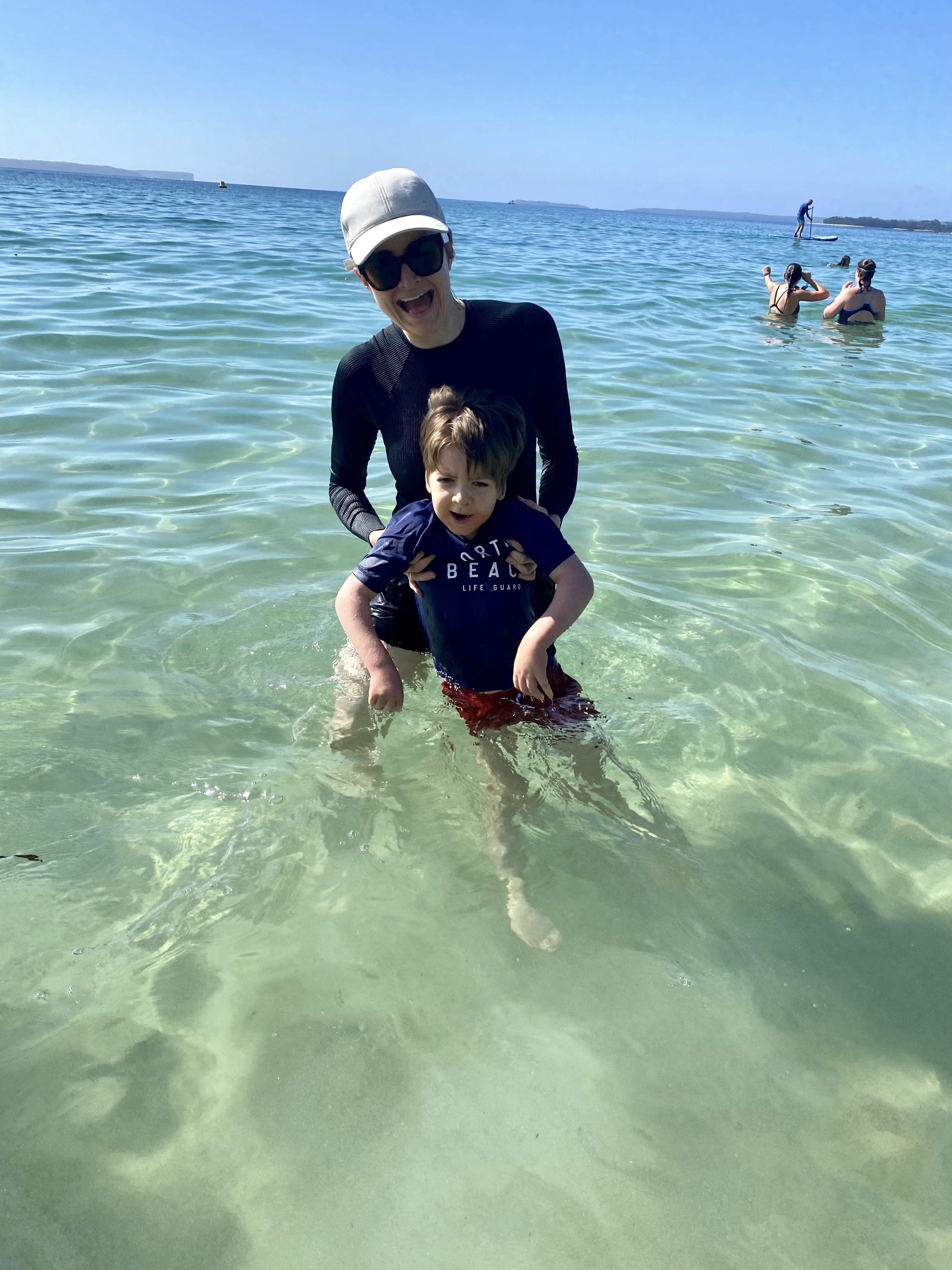 Melanie Dimmitt and her son, Arlo, are swaying in the ocean. Melanie, who is wearing sunglasses, a cap, and a long-sleeved swim shirt, is holding onto Arlo, who is also in swim gear. The sea water is translucent, and other swimmers are nearby.&nbsp;