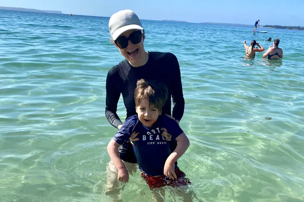 Melanie Dimmitt and her son, Arlo, are swaying in the ocean. Melanie, who is wearing sunglasses, a cap, and a long-sleeved swim shirt, is holding onto Arlo, who is also in swim gear. The sea water is translucent, and other swimmers are nearby.