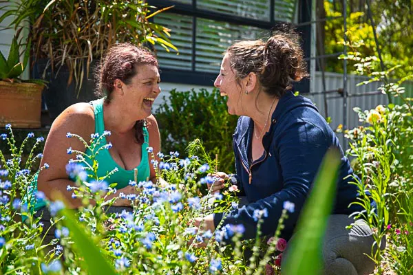 Portia and Sam are knelt down in the garden with flowers all around them. They are staring at each other, laughing uncontrollably.