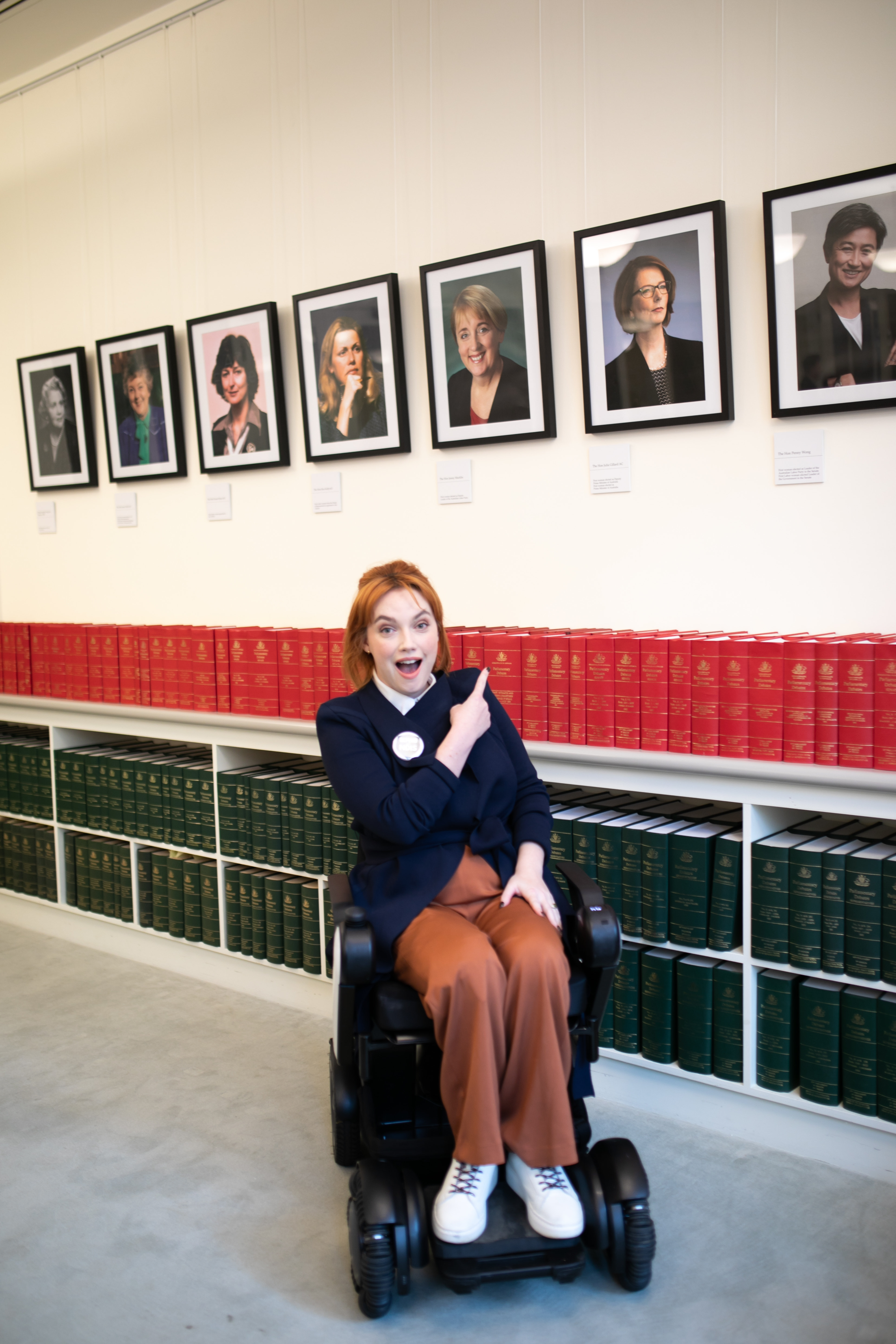 Disability advocate Elly Desmarchelier poses smiling and pointing toward a portrait of former Australian PM, Julia Gillard. Elly has vibrant red hair and wears a navy blazer and rust-coloured pants. She is a wheelchair user and when this photo was taken, was at Parliament house in her role as national spokesperson for the Every Australian Counts 'Save our NDIS' campaign.