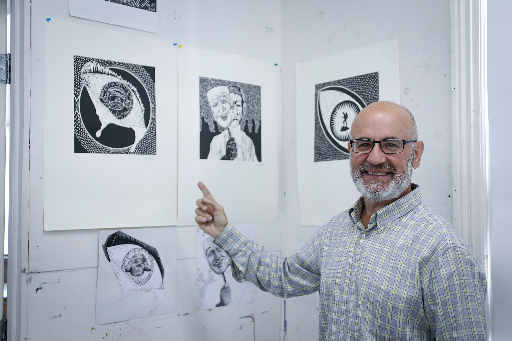 Joe is standing pointing toward his artworks that are hung on the wall.