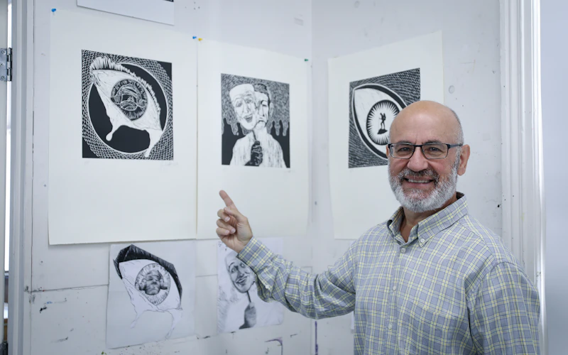 Joe is standing pointing toward his artworks that are hung on the wall.