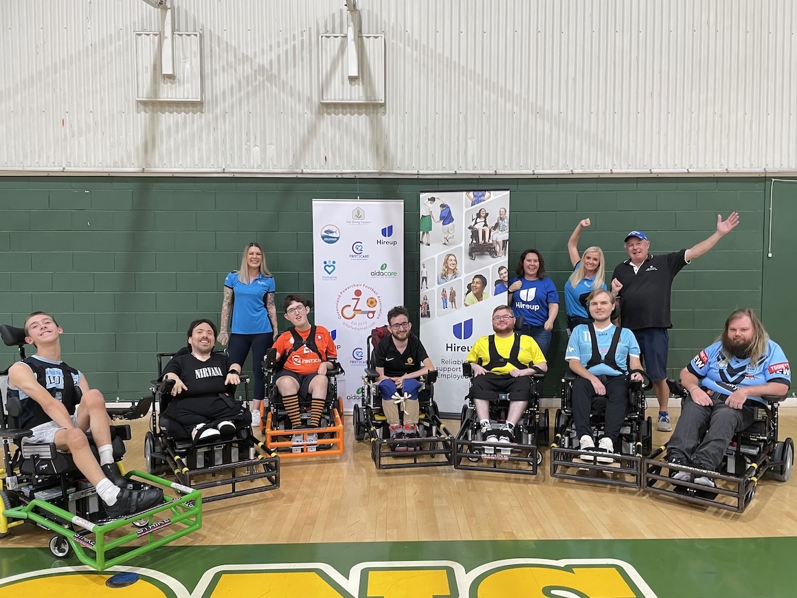 Seven QLD power wheelchair football players sit in power wheelchairs in a semi circle in an indoor court. Four other staff stand in the background. 