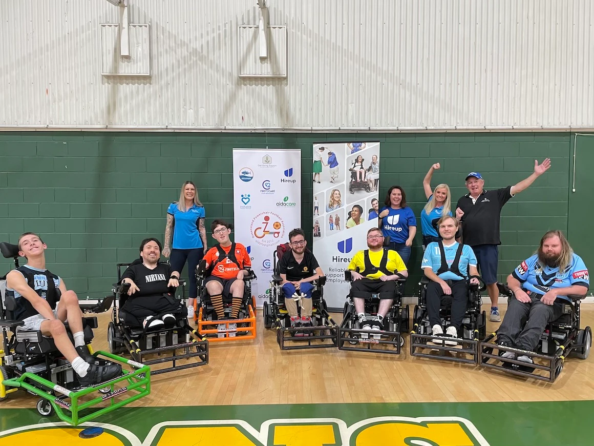 Seven QLD power wheelchair football players sit in power wheelchairs in a semi circle in an indoor court. Four other staff stand in the background.