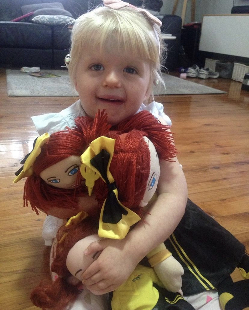 A child with blonde hair and blue eyes cuddles with her three Emma Wiggle toys. 