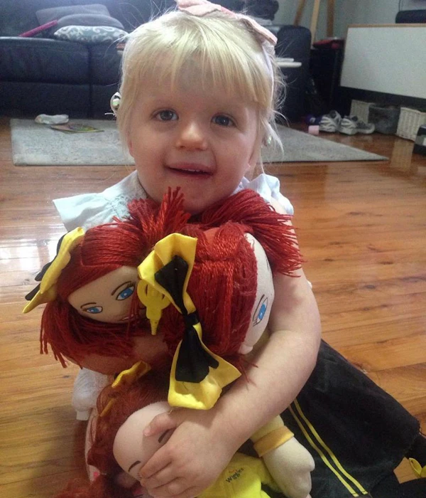 A child with blonde hair and blue eyes cuddles with her three Emma Wiggle toys.