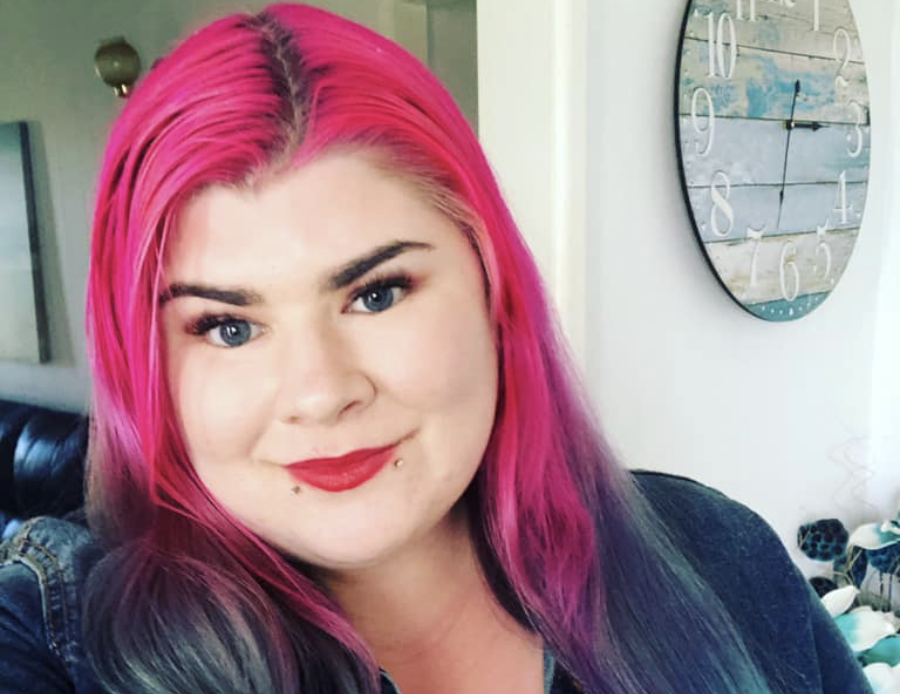 Zoe has long bright pink and aqua hair. She is wearing red lipstick and a blue denim jacket. There is a clock in the background. 