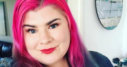 Zoe has long bright pink and aqua hair. She is wearing red lipstick and a blue denim jacket. There is a clock in the background.