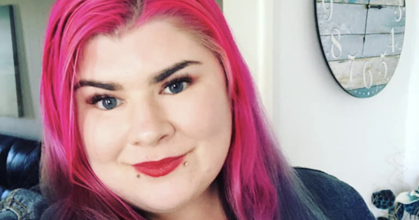 Zoe has long bright pink and aqua hair. She is wearing red lipstick and a blue denim jacket. There is a clock in the background.
