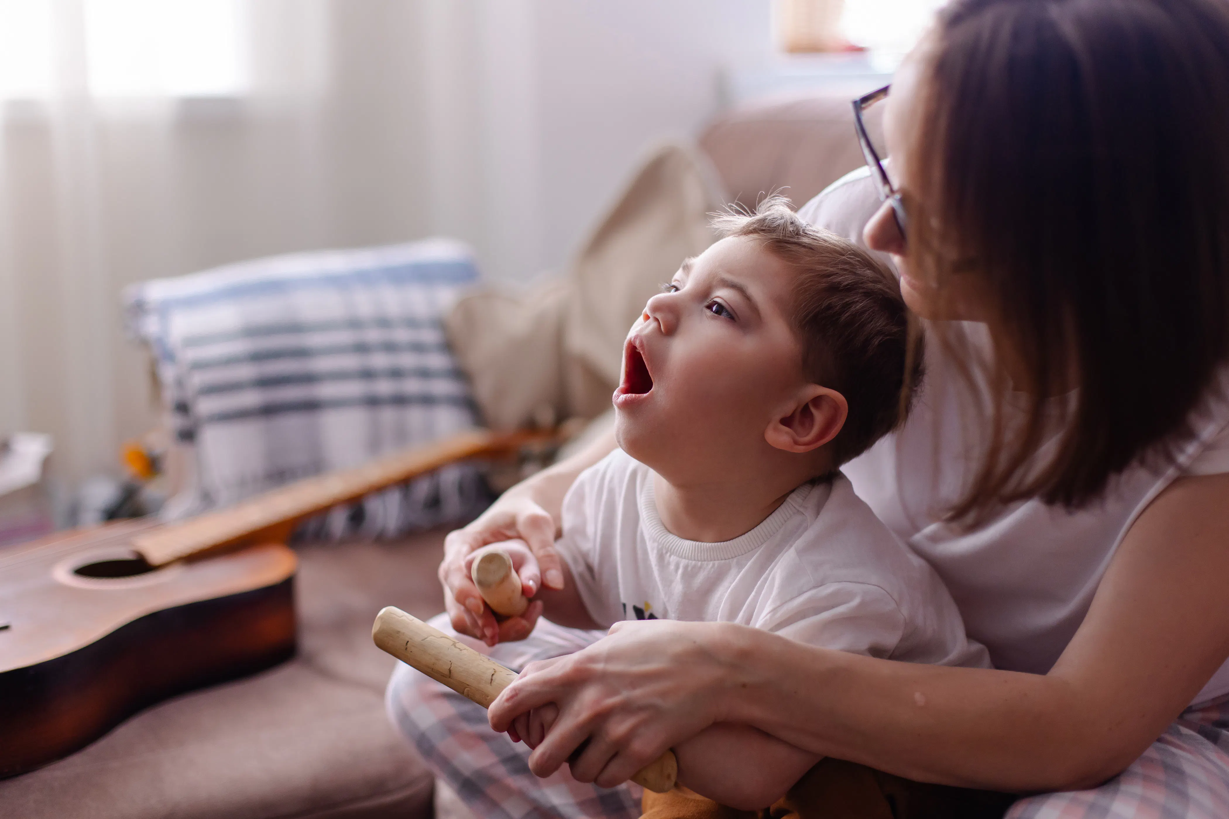A young boy is playing clapping sticks with the help of a woman. He has short brown hair and is wearing a white shirt. There is a brown guitar on a couch in the background.