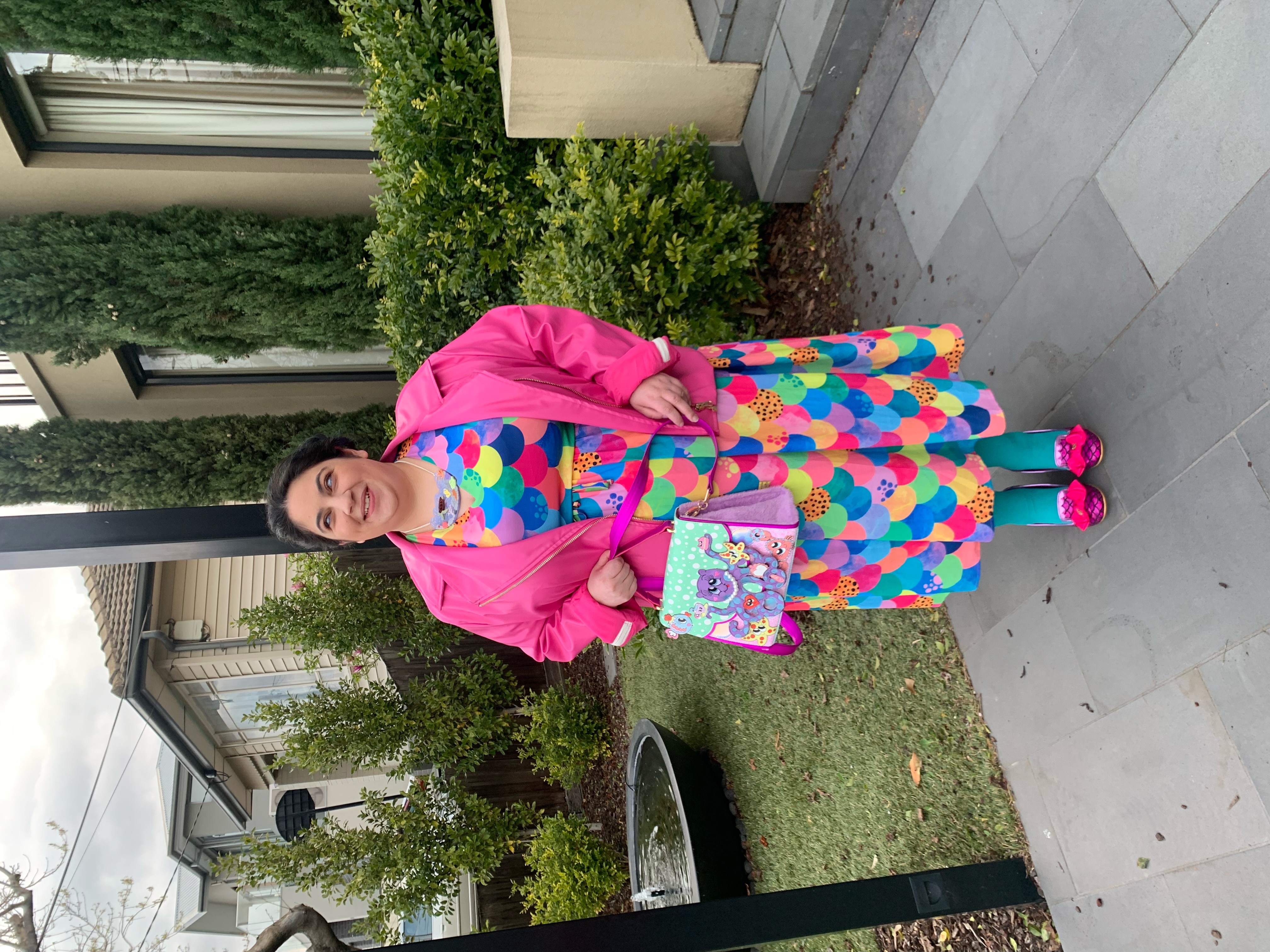 Olivia Muscat, a dark haired, olive skinned woman standing on a path smiling. Olivia is wearing a very colourful dress with a mermaid scale print, pink leather jacket, pink shoes with bows on the toe, a necklace that's a reef scene and a bag featuring an octopus having a tea party.
